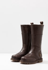 Brązowe skórzane kozaki za kolano z ozdobnymi przeszyciami typu brogue, bocznym zamkiem i uchwytami do zakładania. Solidne podeszwy z fakturowanym wykończeniem.