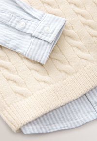 Pull torsadé couleur crème porté sur une chemise à rayures bleues et blanches avec poignets et bas boutonnés.