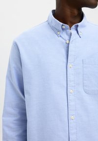 Hombre con una camisa azul claro con botones, bolsillo en el pecho, botones blancos y cuello abotonado, mostrado desde los hombros hasta el cuello.