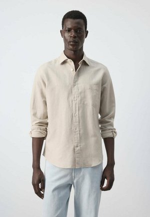 Joven con una camisa beige abotonada con las mangas remangadas y unos vaqueros azul claro, de pie frente a un fondo claro y sencillo.