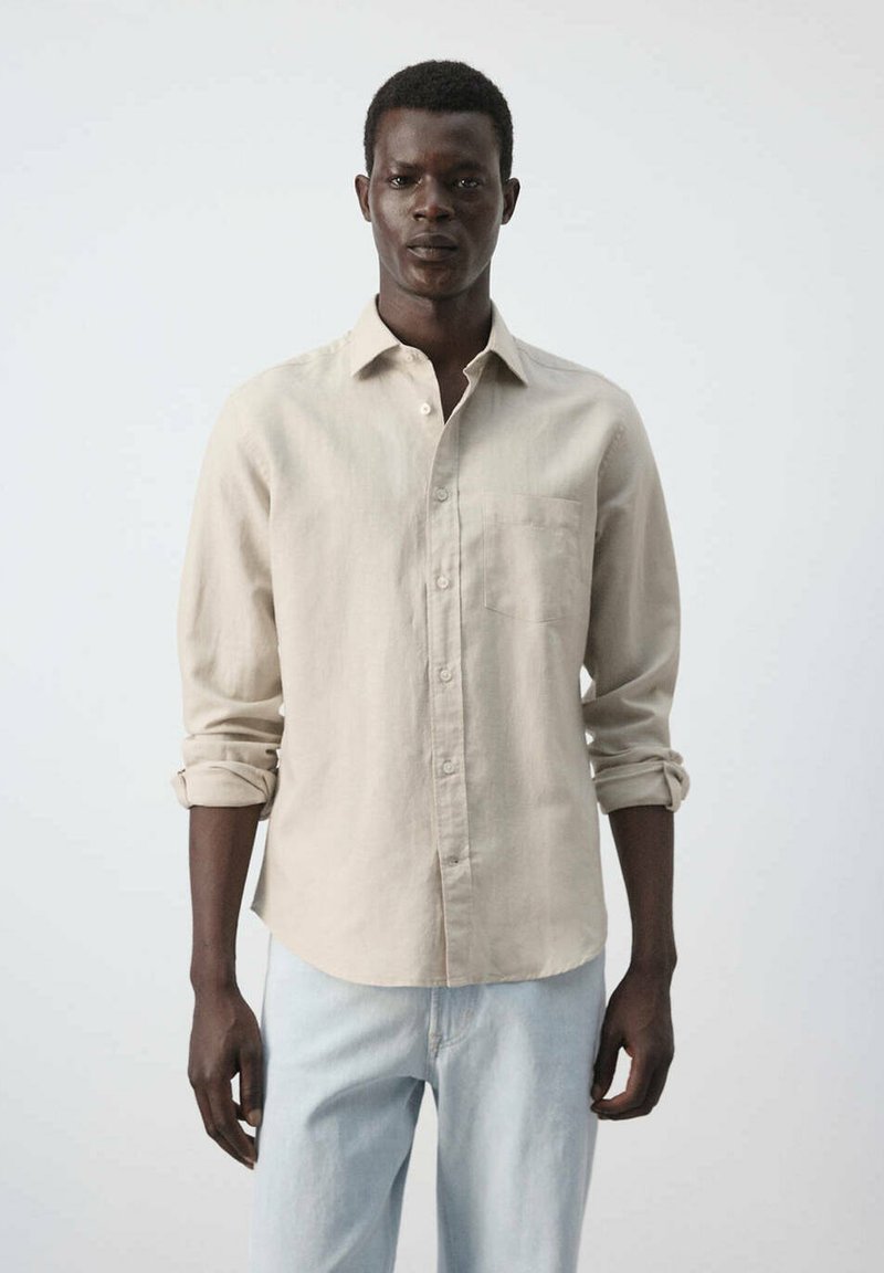 Jeune homme portant une chemise beige à boutons avec les manches retroussées et un jean bleu clair, debout devant un fond clair uni.