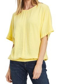 Żółta lniana bluzka z krótkim rękawem, okrągłym dekoltem i zebranym wykończeniem, w połączeniu z ciemnymi dżinsami. Miękka tekstura i luźny krój.