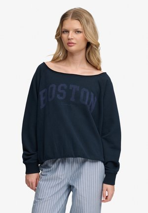 Jeune femme aux longs cheveux blonds portant un sweat-shirt bleu marine à épaules dénudées avec le texte "BOSTON" et un pantalon gris clair à fines rayures.