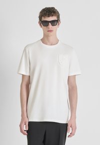 Camiseta de algodón blanca de mangas cortas, con un bolsillo texturizado que presenta un diseño de escudo. Se lleva con gafas de sol rectangulares negras.