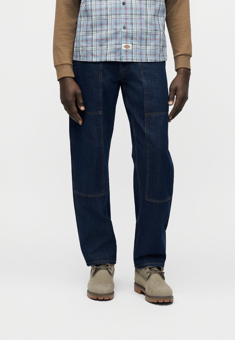 Mann trägt dunkelblaue Jeans mit geradem Bein, beige Stiefel, ein tanfarbenes Langarmshirt und darüber ein blau kariertes Hemd zum Knöpfen.