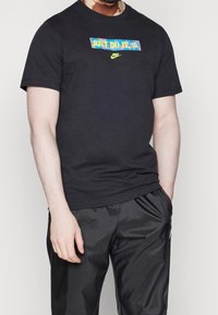 Musta puuvillapaita, jossa on värikäs "Just Do It!" -grafiikka ja pieni Nike-logo keskellä. Lyhyet hihat ja pyöreä kaula-aukko.