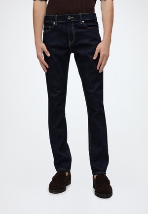 Homme portant un jean slim bleu foncé et des mocassins marron foncé, debout sur fond blanc.