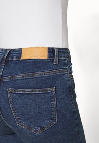 Tmavě modré džíny z denimu s koženou značkovou nášivkou označenou "NOISY MAY." Mají standardní kapsy a kontrastní švy. Přiléhavý střih.