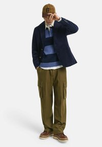 Corduroy blazer in navy, striped blue sweater, white shirt, olive cargo pants with side pockets, brown shoes, and patterned cap with logo.