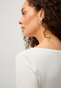 Silberne Creolen werden von einer Person mit lockigem Haar getragen. Das hellfarbige Oberteil hat einen runden Halsausschnitt, und Haut ist am Hals und im Gesicht sichtbar.