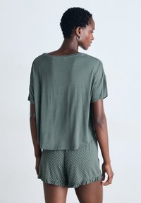 Kvinna med kort lockigt hår klädd i en grön oversize t-shirt och gröna shorts med prickar och flikiga kanter, vänd framåt mot en vit bakgrund.