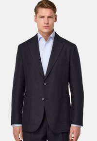 Blazer en lin marine foncé avec deux boutons, revers notches et deux poches avant. Porté sur une chemise rayée bleu clair. Coupe ajustée.