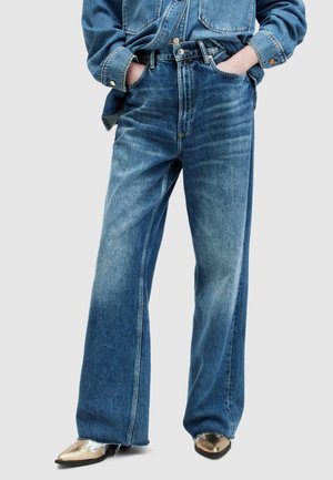 Jeans de mezclilla azul de pierna ancha con un acabado desgastado, cintura alta y ligero distress en el dobladillo, combinados con zapatos de punta metálica dorada.