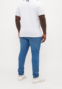 Bílý tričko s krátkým rukávem a kulatým výstřihem, spárované s modrými džíny a bílými teniskami s šedými akcenty. Pohled zezadu, jednobarevné pozadí.