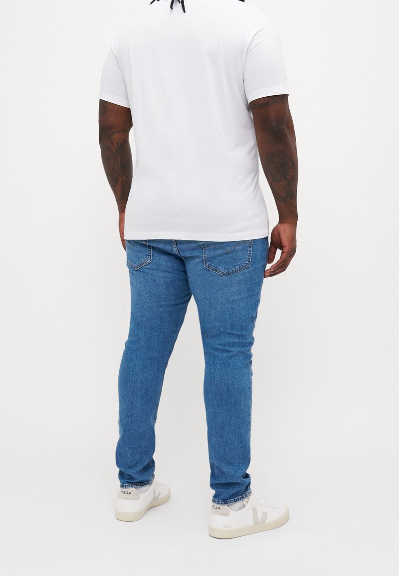 Bílý tričko s krátkým rukávem a kulatým výstřihem, spárované s modrými džíny a bílými teniskami s šedými akcenty. Pohled zezadu, jednobarevné pozadí.