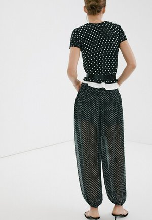 Žena u crnoj polka dot majici kratkih rukava i širokim prozirnim hlačama na točkice, stoji s rukama iza leđa ispred jednostavne pozadine.