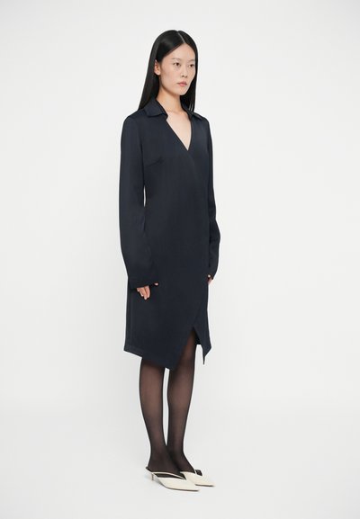 Rochie închisă navy, tip wrap, cu mâneci lungi și decolteu adânc în V, având un buzunar aplicat și un tiv care se oprește deasupra genunchiului. Purtați cu mule albe.