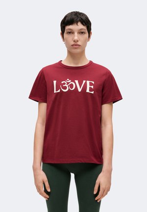 Persona con cabello corto y oscuro que lleva una camiseta roja con texto blanco "LOVE" que presenta un símbolo Om en lugar de la letra O, y pantalones verde oscuro.