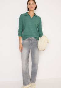 Frau in grünem Strickpullover und ausgebleichten grauen Jeans, die eine weiße gepolsterte Jacke vor einem schlichten weißen Hintergrund hält.