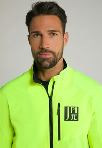 Hochsichtbare gelbe Jacke mit schwarzen Akzenten, durchgehendem Reißverschluss und einer Brusttasche mit Reißverschluss, die auf der linken Seite ein Logo trägt. Glatte Textur.