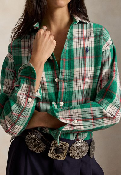 Femme portant une chemise à carreaux verte et blanche boutonnée, ceinture concho argentée et pantalon sombre, main près du col.