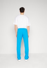 Man som står vänd bort med en vit t-shirt, ljusblå träningsbyxor och vita sneakers mot en enkel ljusgrå bakgrund.