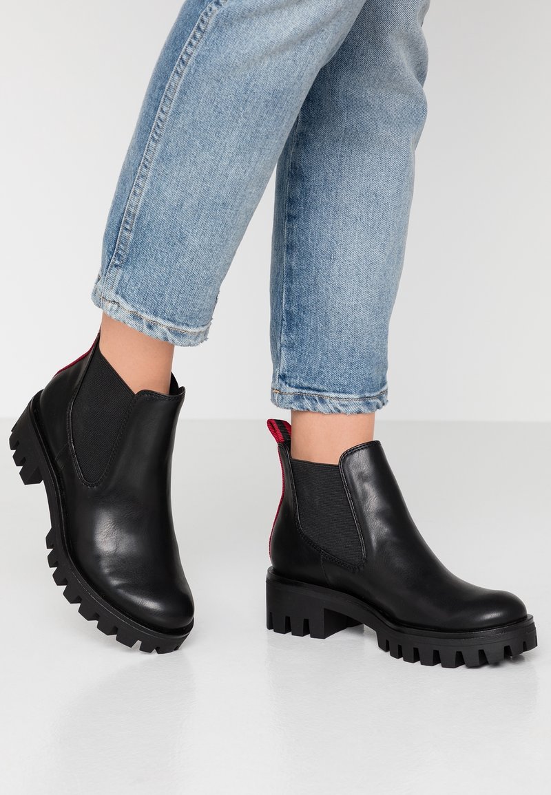 Bottes courtes en cuir noir avec semelles crantées épaisses, languettes rouges, portées avec des jeans bleu clair retroussés sur fond blanc.