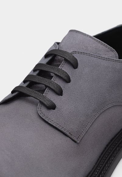 Chaussure habillée en daim gris avec des lacets noirs texturés et des coutures visibles sur le dessus et près de la semelle.