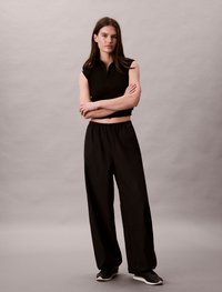 Μαύρη αμάνικη μπλούζα με φερμουάρ στον γιακά, συνδυασμένη με χαλαρούς μαύρους παντελόνες, τονισμένες με ευρύ κόψιμο και ελαστική μέση. Μαύρα αθλητικά παπούτσια.