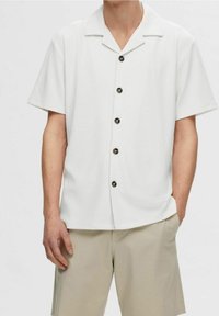 Man wearing a short-sleeve white button-up shirt and beige shorts with one hand in pocket against a plain background.