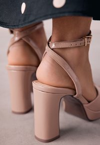 Bézs platform magassarkú fényes kivitelben, boka pánttal és vastag sarokkal. A dizájn keresztpántokat és sima felületet tartalmaz.