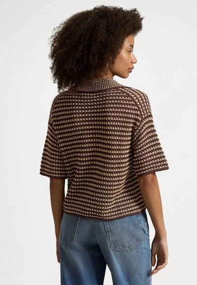 Ženska s kodrastimi lasmi, oblečena v kratko majico z rjavimi in bež črtami iz pletenine ter modre kavbojke, prikazana od zadaj.