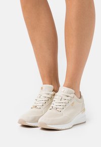 Béžové sportovní boty s texturovaným síťovaným svrškem, plochými tkaničkami a bílou podešví. Zlaté akcenty na dírkách na tkaničky a bočním detailu.