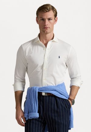 Weißes Langarmhemd, marineblau gestreifte Hose, blauer Pullover um die Taille gebunden, klassischer Kragen und dezentes Logo-Detail auf dem Hemd.