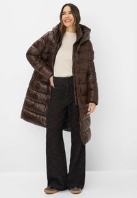 Brun pufferjakke med en skinnende overflade, hætte og integreret lynlås; båret over en beige ribbet sweater og teksturerede sorte breddebenbukser.