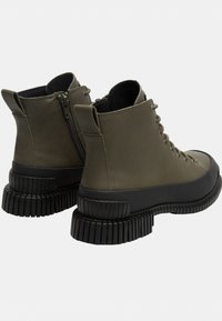 Botas de tobillo de cuero verde oliva con suelas de goma negras acanaladas, con cordones en la parte delantera y cremallera lateral para el cierre.