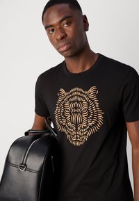 Black cotton t-shirt featuring a large gold tiger face graphic. Model holds a black leather backpack with a silver zipper.