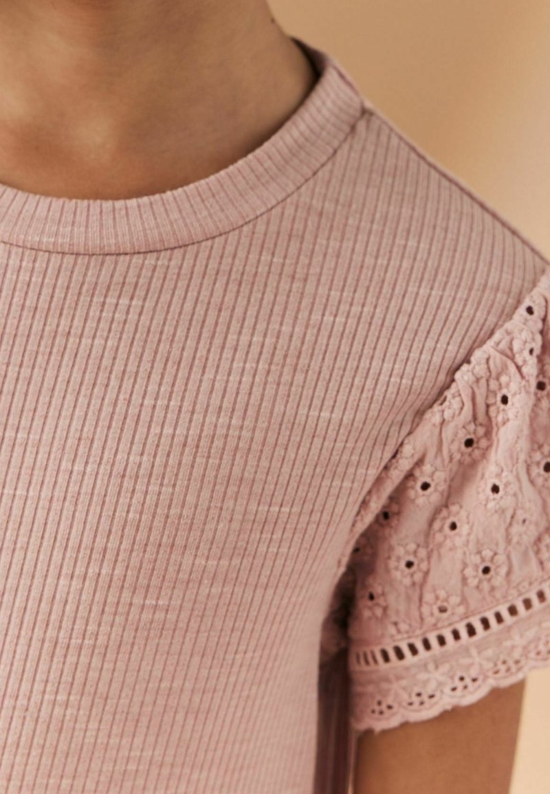 Gros plan d'une personne portant un haut côtelé rose clair avec de courtes manches en dentelle décorées de broderies florales et de petites découpes.