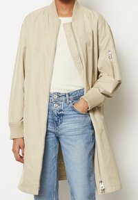 Lichtbeige bomberjack met ribgebreide manchetten en kraag, ritssluiting aan de voorkant en zijzak, gecombineerd met lichtblauwe jeans en een witte ribgebreide top.