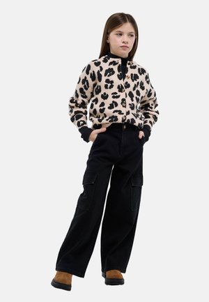 Fille aux cheveux bruns raides portant un cardigan imprimé léopard, un pantalon cargo noir à jambes larges et des bottes marron, debout les mains dans les poches.