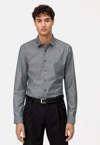 Camicia grigia con finitura testurizzata e colletto a punta, dotata di una tasca sul petto e maniche lunghe, abbinata a pantaloni neri e cintura.