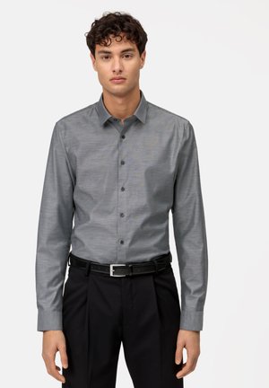 Camicia grigia con finitura testurizzata e colletto a punta, dotata di una tasca sul petto e maniche lunghe, abbinata a pantaloni neri e cintura.