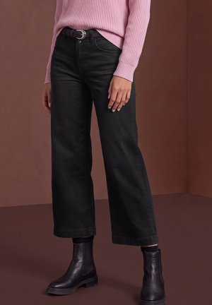 Le mannequin porte un jean noir à jambes larges avec une ceinture, un pull en maille rose et des bottines noires.