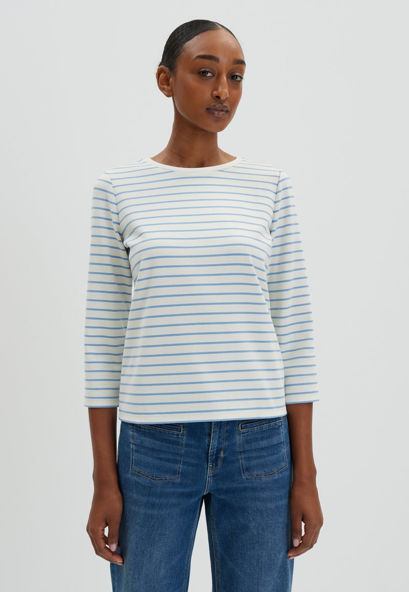 Weißes Langarmoberteil mit hellblauen horizontalen Streifen, runder Halsauschnitt und weichem Stoff. Kombiniert mit blauen Jeans.