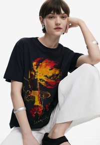 Schwarzes T-Shirt mit buntem Blumenmotiv, kurze Ärmel, lockere Passform. Das Modell trägt ein silbernes Armreif und posiert mit weißen Hosen.