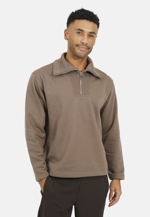 Uomo con capelli corti e ricci che indossa un pullover taupe a mezza zip e pantaloni scuri, in piedi con una mano in tasca su uno sfondo bianco.