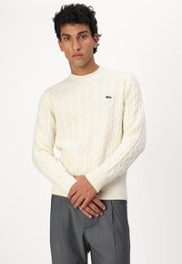 Cream cable-knit sweater with ribbed cuffs and hem; features a small green logo on the chest. Paired with gray trousers.