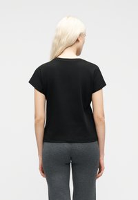 Γυναίκα με μακριά ξανθά μαλλιά, φορώντας μαύρο κοντομάνικο μπλουζάκι και γκρι παντελόνι, στέκεται με το σώμα γυρισμένο προς τα πίσω μπροστά σε απλό φωτεινό φόντο.