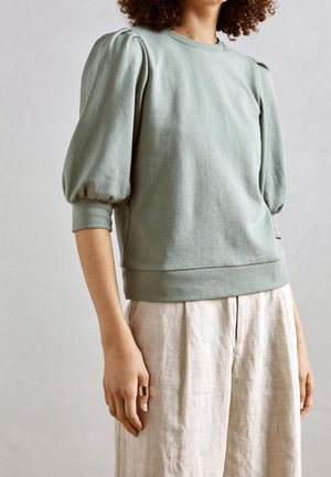 Kobieta w zielonym topie z bufiastymi rękawami i luźnych beżowych spodniach, z złotym pierścieniem na lewej dłoni na tle jednolitego jasnego tła.