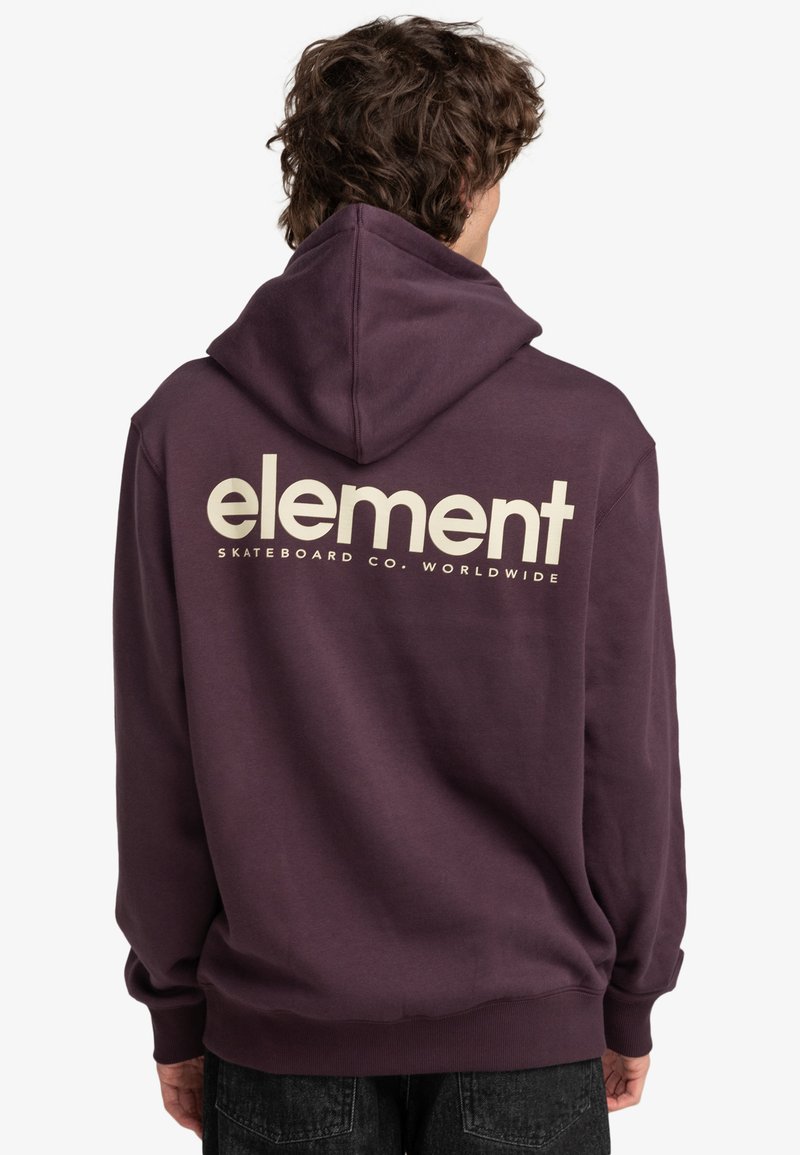 Persona con cabello rizado vistiendo una sudadera con capucha de color púrpura oscuro que lleva el texto "element Skateboard Co. Worldwide" en la parte posterior, combinada con jeans negros.
